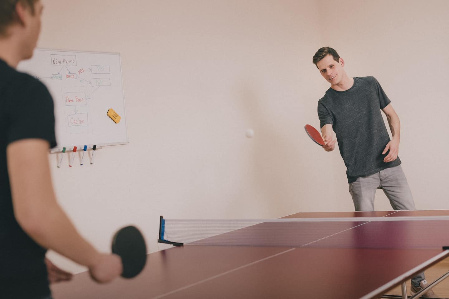 Dvaja muži hrajú stolný tenis v kancelárii s bielou tabuľou v pozadí.