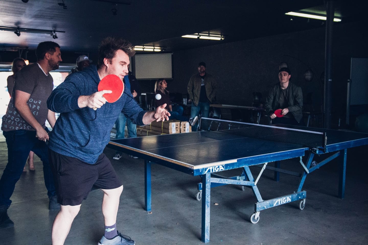Muž v modrej mikine hrá stolný tenis s kamarátom.